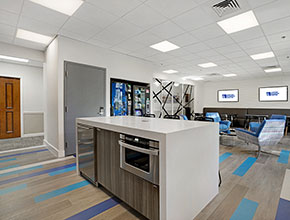 A break room with a kitchen island with high chairs, some vending machines in the wall, and numerous chairs and tables